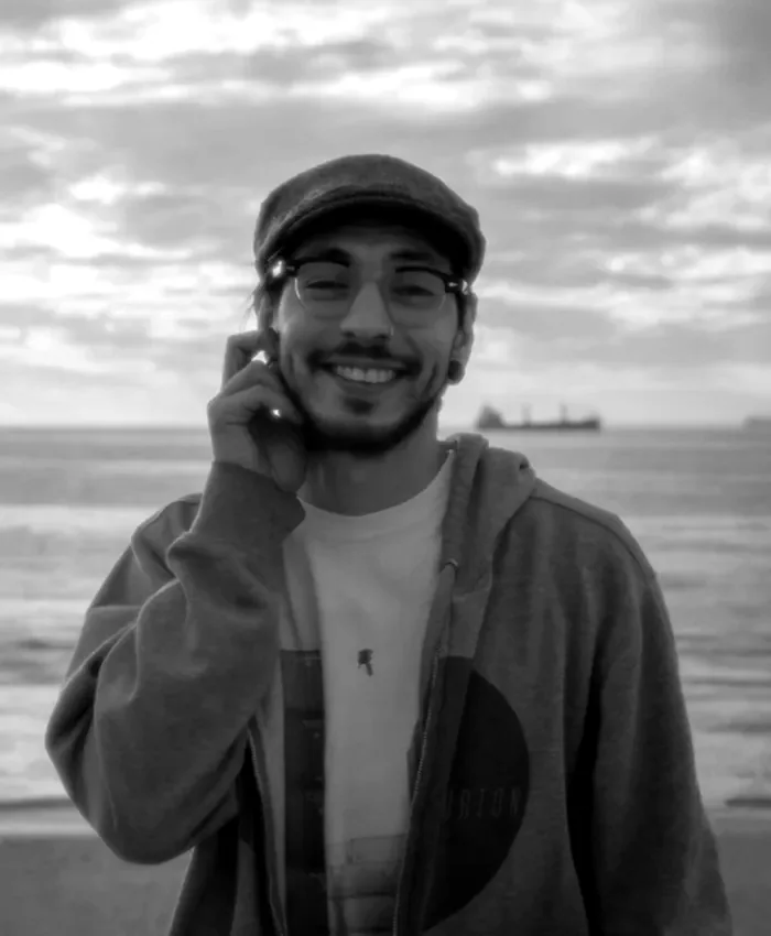 Pablo, bajo y coros, sonriendo con gorra y lentes frente al mar